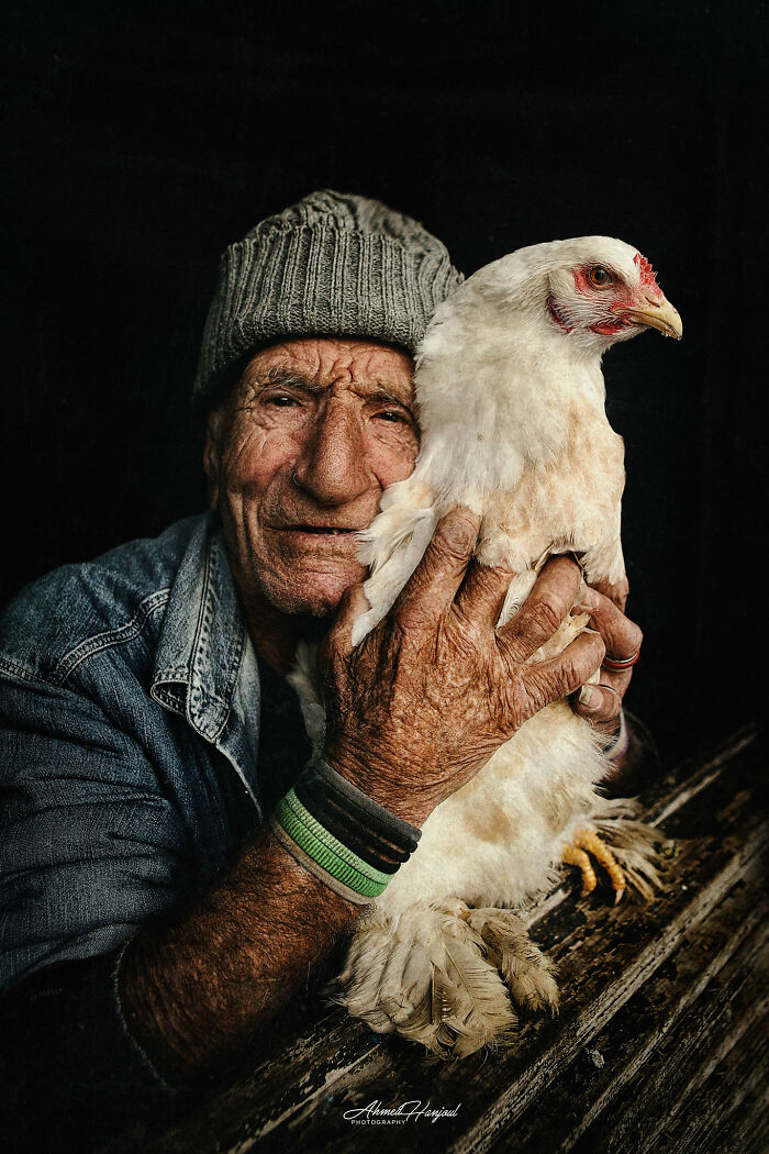 Photographer captures expressive face of an elderly man in a beanie, holding a white chicken, against a dark background.
