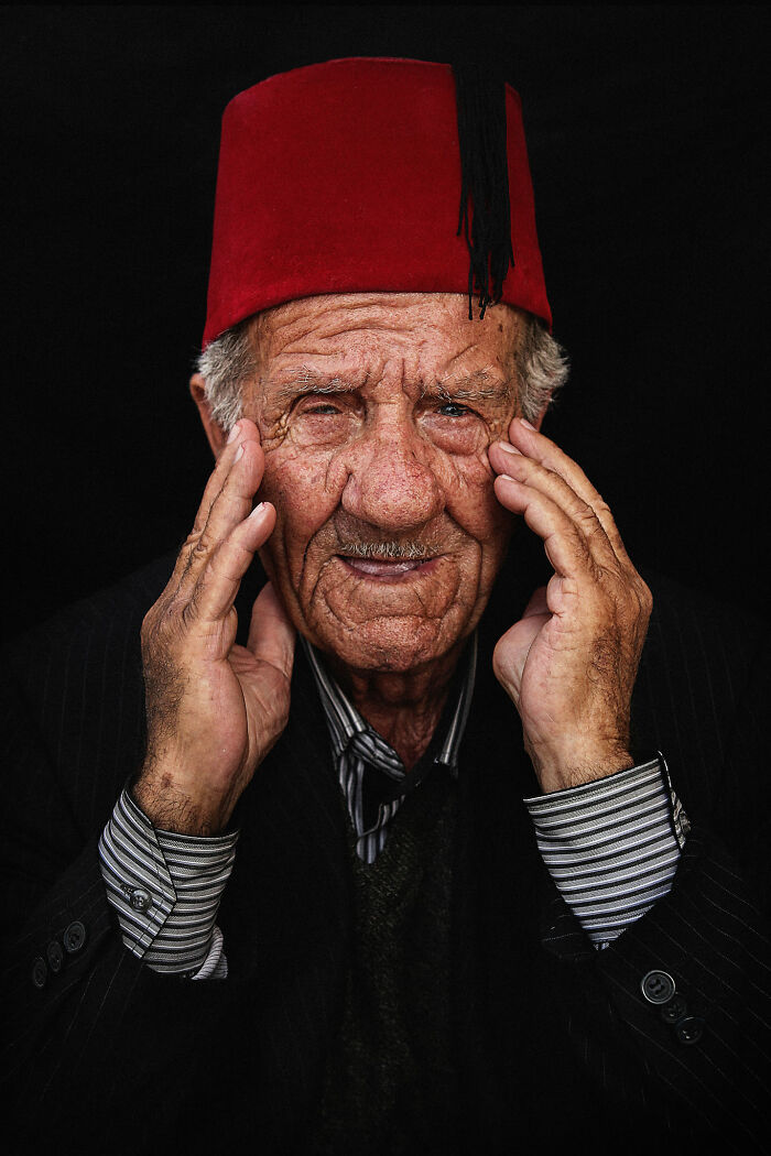 Elderly man wearing a red hat, with expressive face and hands on cheeks, photographed by a talented photographer.
