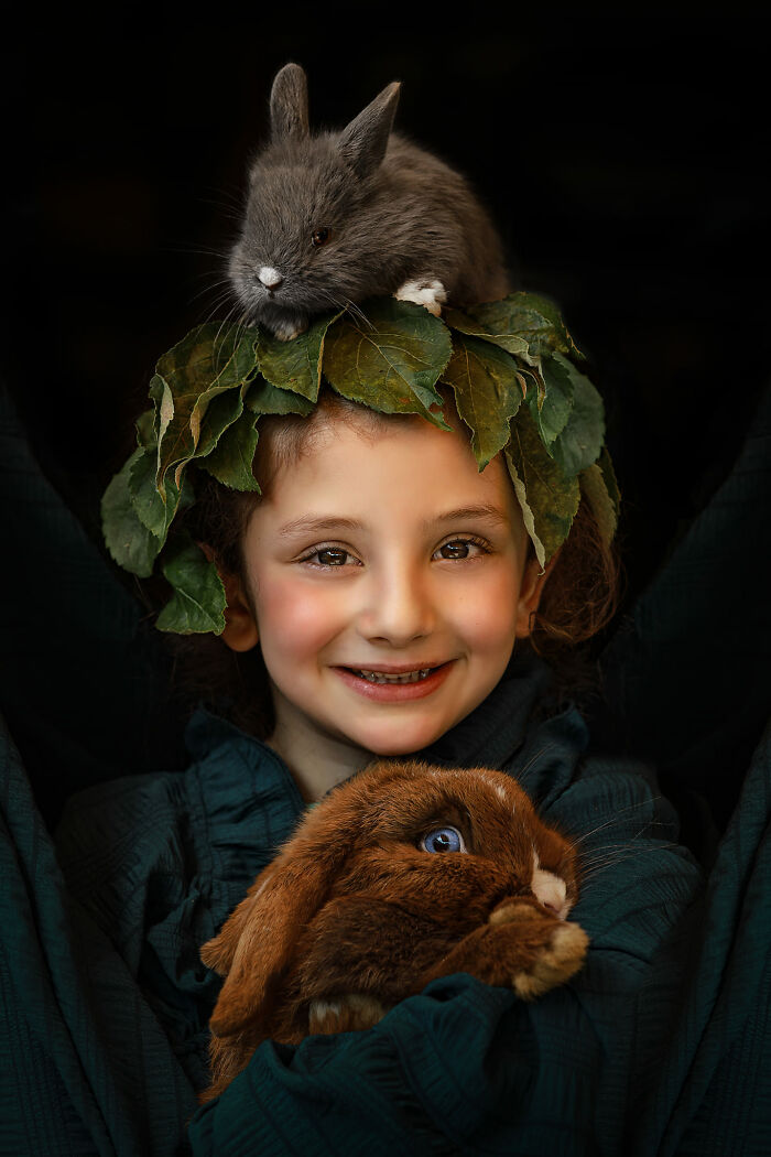 A child with expressive face holds a rabbit, another rabbit on head, wearing leaf crown, creating a storytelling portrait.