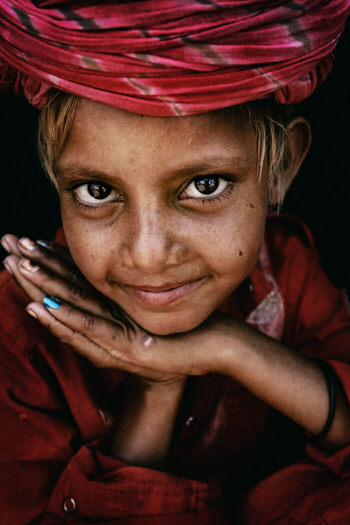 Expressive face of a child with a colorful headscarf, hands under chin, captured by photographer.