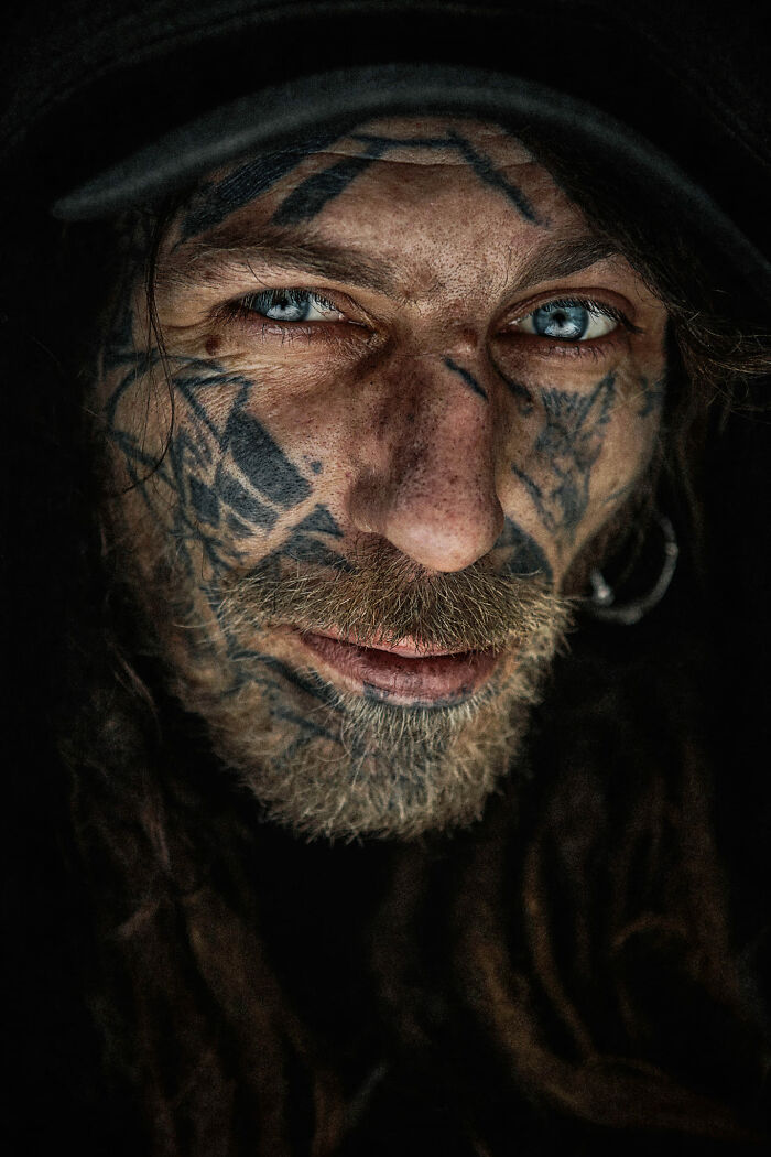 Expressive face with tattoos, intense blue eyes, and a textured beard captured by a photographer.