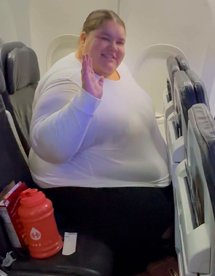 Passenger seated on a plane, waving and smiling, surrounded by travel items, amidst debate on airline discrimination fees.