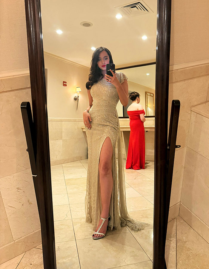 Woman in an elegant gold dress taking a mirror selfie in a restroom, with another woman in red in the background. Woman in an elegant gold dress taking a mirror selfie in a restroom, with another woman in red in the background.