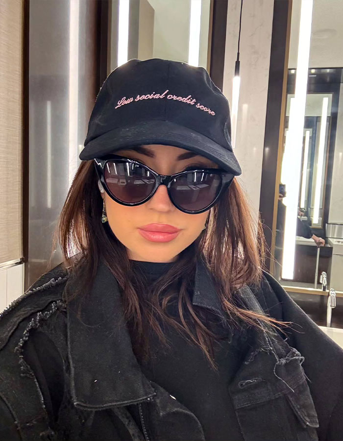 Woman in sunglasses and black cap with text, taking a selfie indoors; SEO keywords: Ashley St. Clair. Woman in sunglasses and black cap with text, taking a selfie indoors; SEO keywords: Ashley St. Clair.