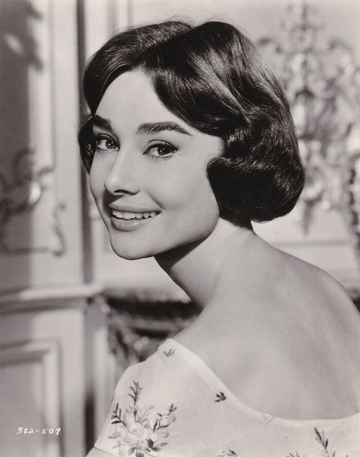 Black and white portrait of a smiling celebrity with vintage hairstyle, wearing an elegant floral dress.