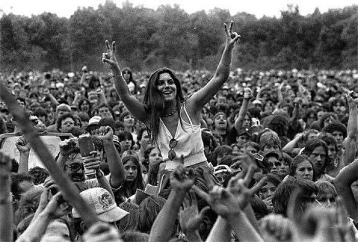 Crowd at a music festival, a woman smiling and raising peace signs, capturing true happiness amidst a sea of people.