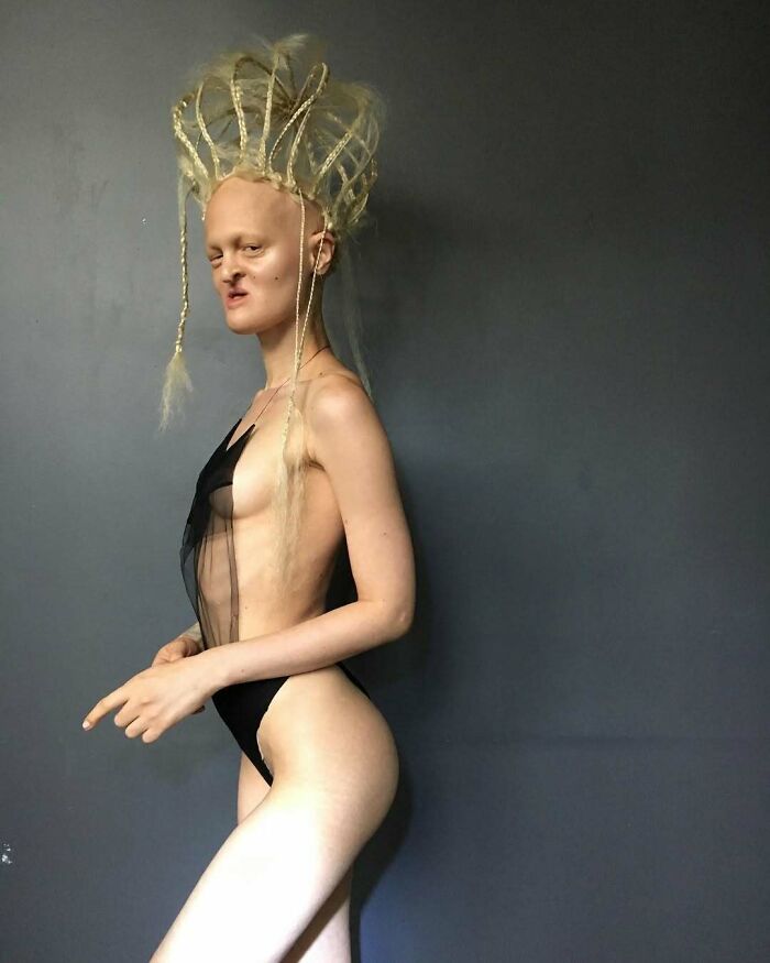 Unique model posing in profile, showcasing avant-garde fashion with an intricate hairstyle against a dark backdrop.