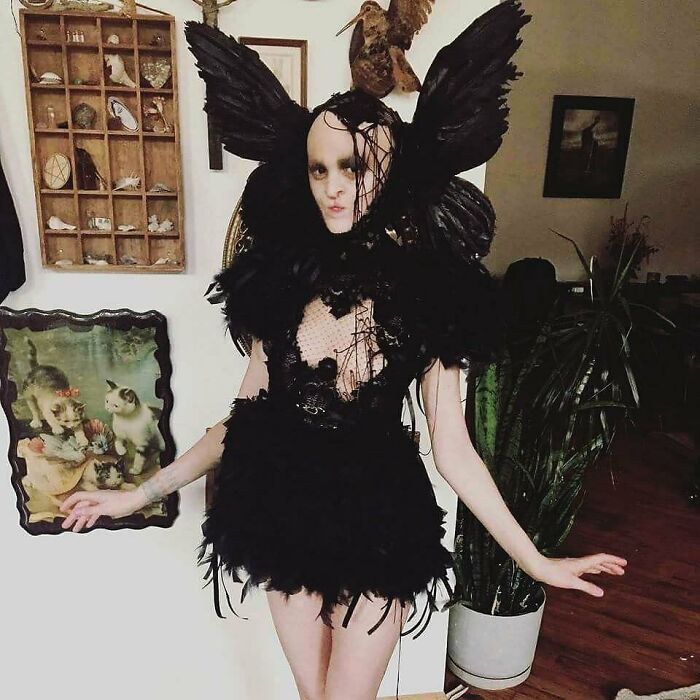 Unique model in black feathered outfit posing in an eclectic room with artwork and plants.