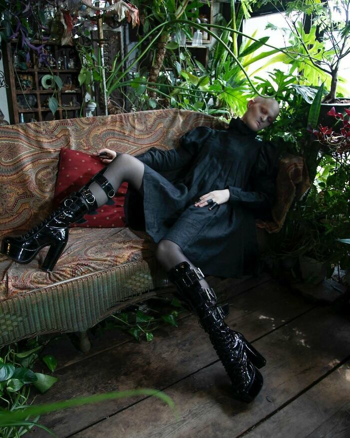 Unique model in black dress and glossy boots, posing on a patterned sofa amidst lush greenery.