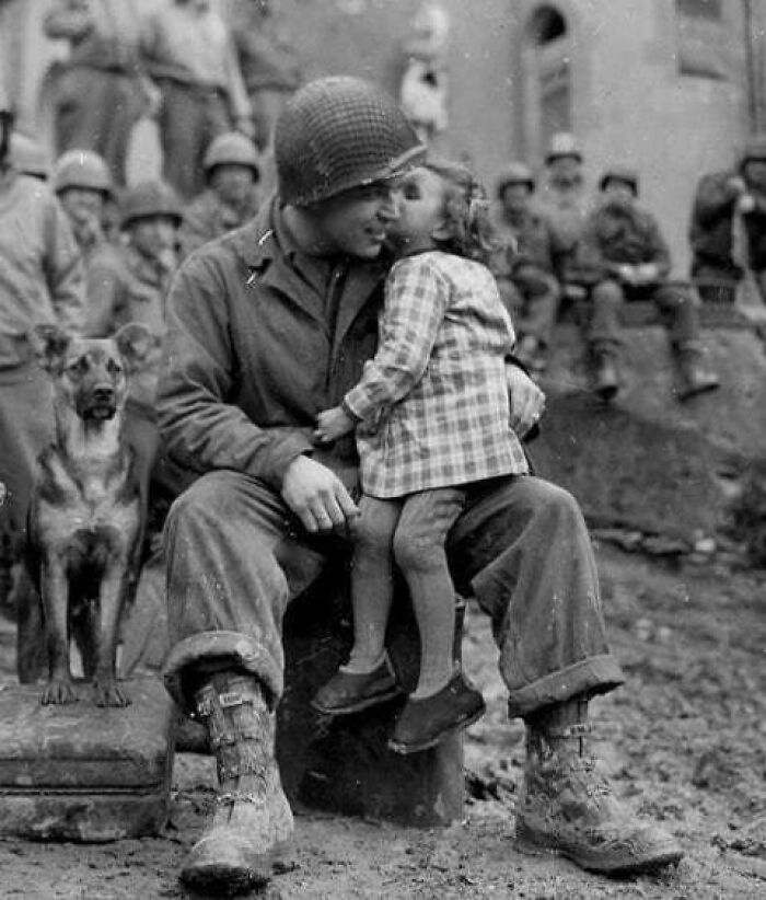 Soldier smiling as child gives a kiss, capturing true happiness; dog sits beside them, surrounded by other soldiers.