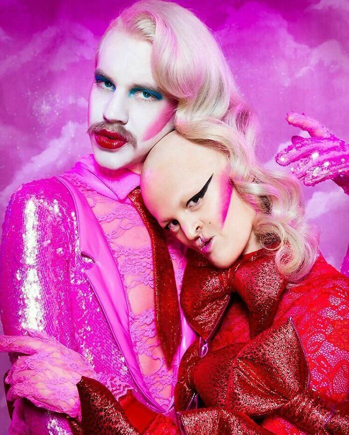 Unique model in vibrant pink and red makeup, striking a bold pose against a dreamy pink background.