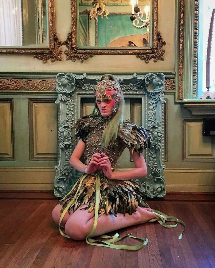 Unique model in ornate feather outfit kneeling in an opulent room with vintage decor and mirrors.