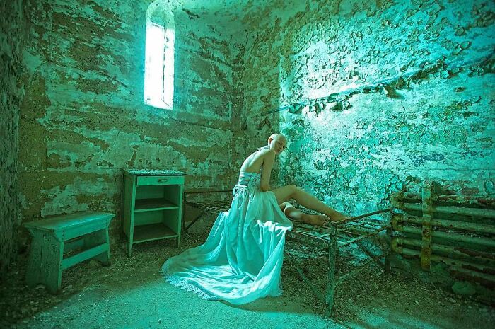 Unique model in a textured room with ethereal lighting, draped in a flowing gown, evoking a surreal atmosphere.