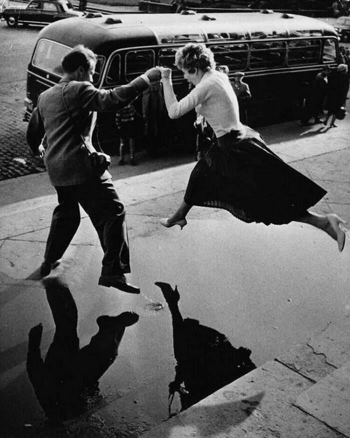 A joyful couple leaps over a puddle, capturing true happiness against a backdrop of a vintage bus and city street.