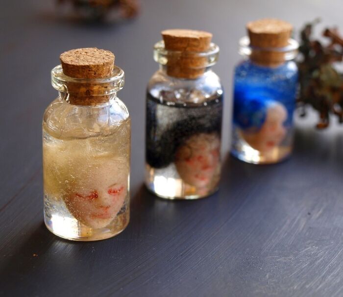 Three creepy handmade crafts in jars with doll heads, filled with colored liquids, on a dark surface.