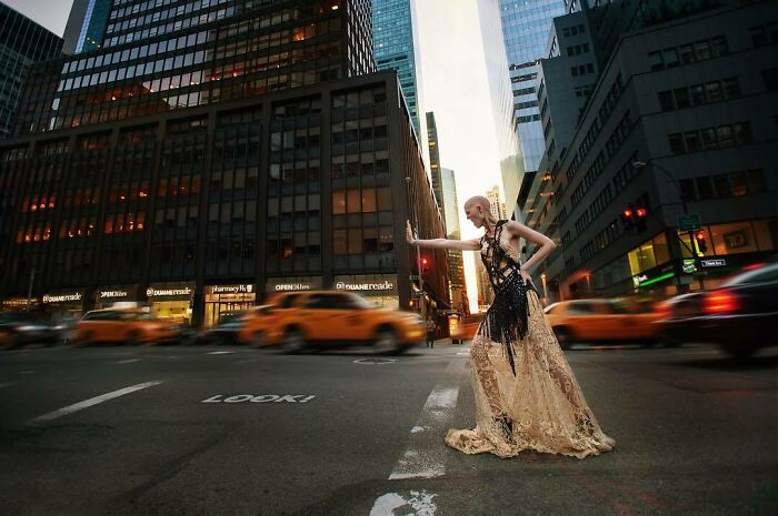 A unique model in an intricate gown stands poised on a busy city street with blurred yellow taxis in the background.