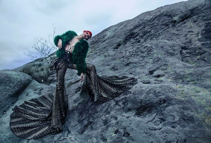 Unique model posing in artistic attire on rocky terrain, wearing a green fur coat and patterned flared pants.