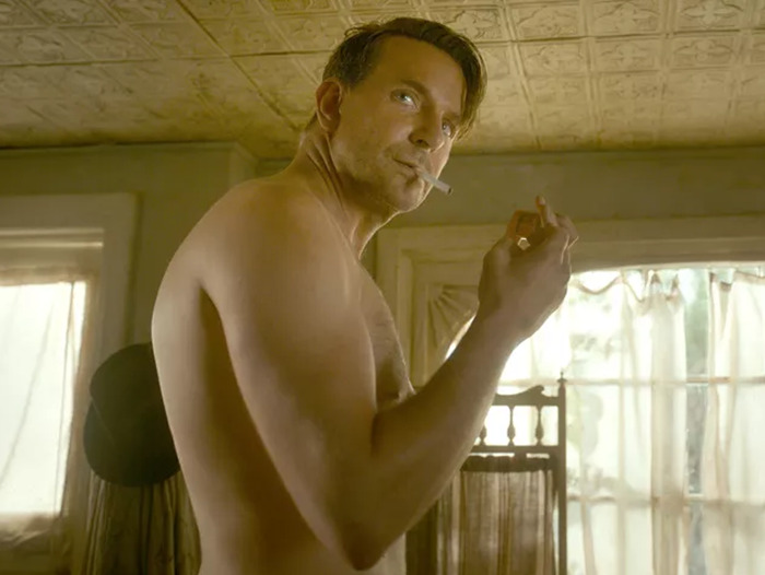 Bradley Cooper in a room, shirtless, holding a cigarette, embodying natural performance without prosthetics.