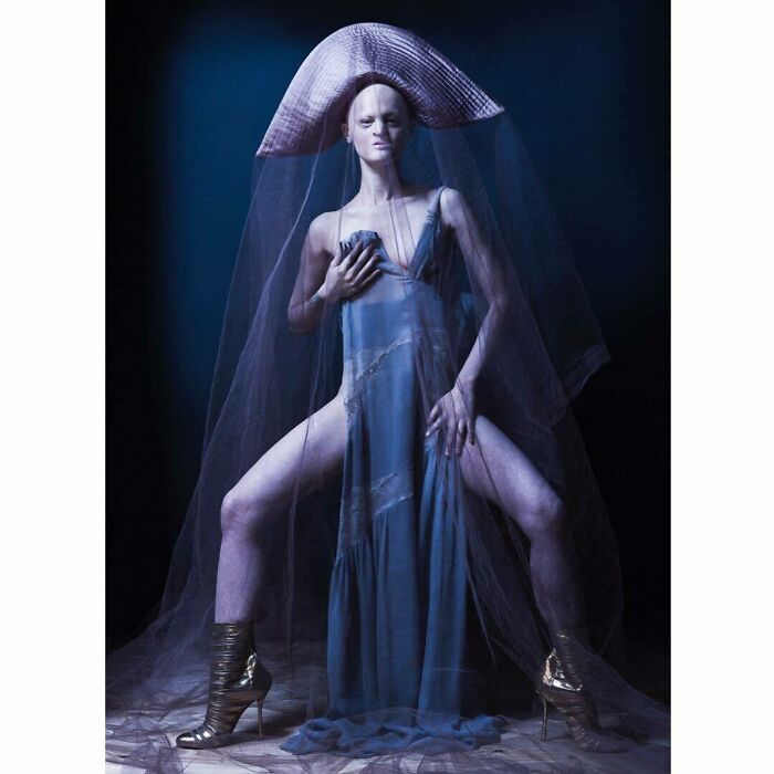 Unique model in avant-garde fashion, wearing a veil and blue dress with gold heels, posing in a dramatic setting.