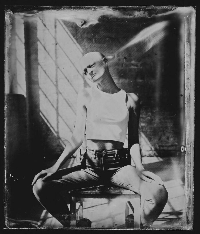 Unique model in a black-and-white photo sitting on a stool, wearing a white top and jeans, with dramatic lighting.