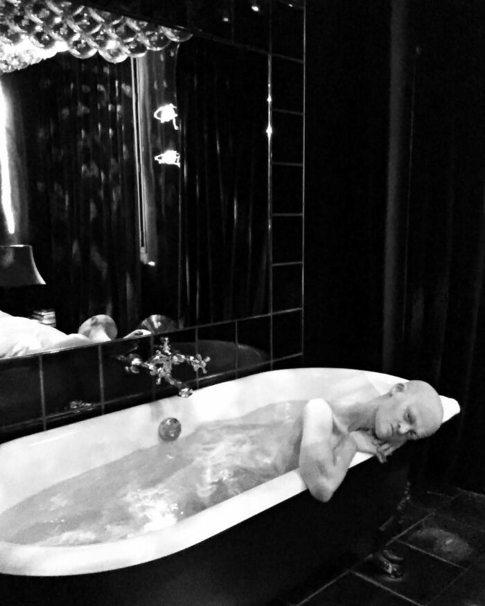 Unique model relaxing in a vintage bathtub, surrounded by dark, reflective surfaces.