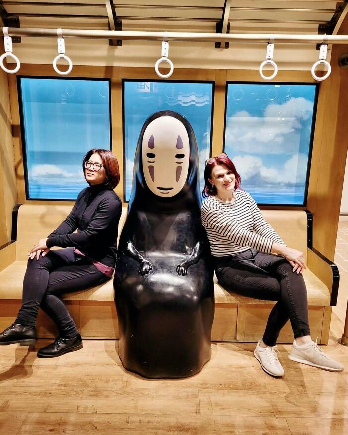 Two people sit beside a No-Face statue on a Japanese train-themed bench. Interesting-Japan-Pics.