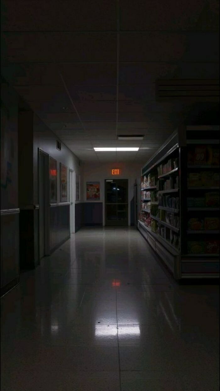 Dimly lit liminal space in a supermarket aisle with an eerie atmosphere and a glowing exit sign.