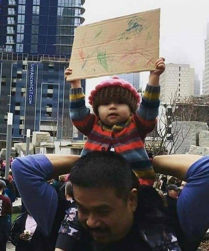 Niña pequeña en hombros sosteniendo un cartel en ciudad, ejemplo divertido de preferiría que no.