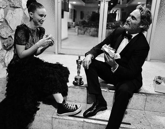 A couple in formal attire sit happily on stairs, sharing snacks by an award statue.