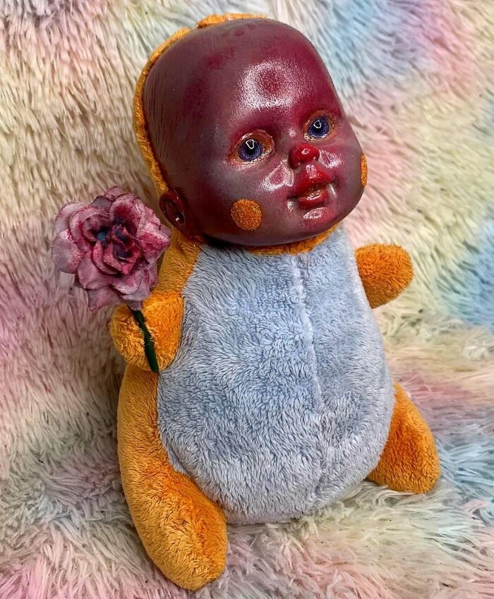 Creepy handmade craft of a baby doll with a painted face in a fuzzy costume holding a fabric rose.