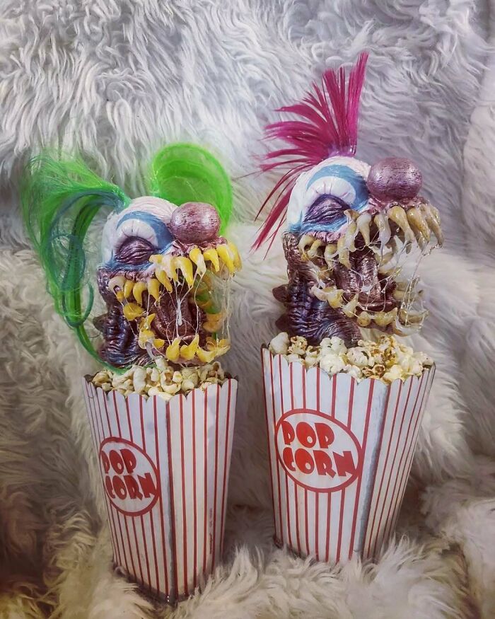 Creepy handmade crafts featuring grotesque clown faces with colorful hair, emerging from popcorn containers on a fluffy backdrop.