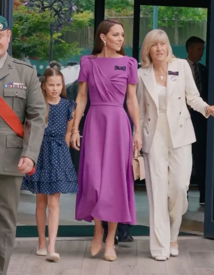 Kate Middleton in a purple dress, walking with others, embodying her fashion policy amidst public attention.