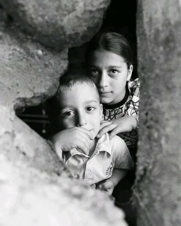 Black and white photograph of two children peering through a stone wall, capturing childhood's innocence and resilience.