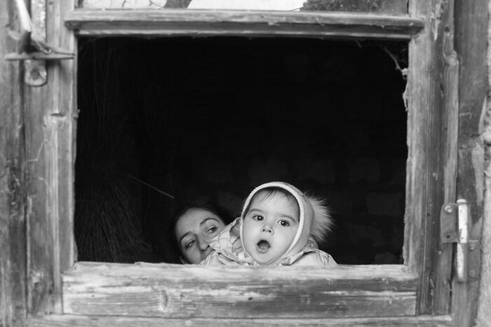 Black and white photo of a baby in a hooded coat, peering out of a rustic window, symbolizing childhood innocence and resilience.