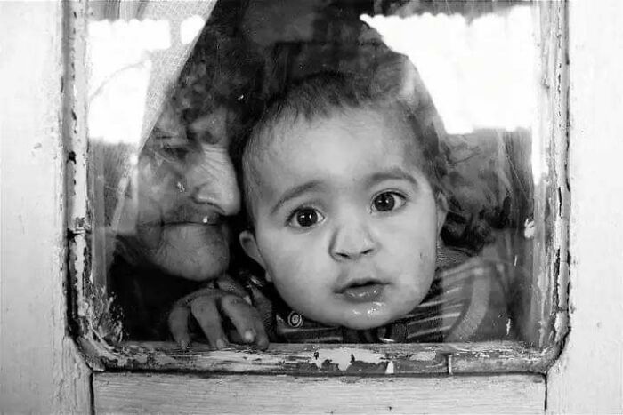 Black and white photo of a child and older woman peering through a window, showcasing childhood's innocence and resilience.