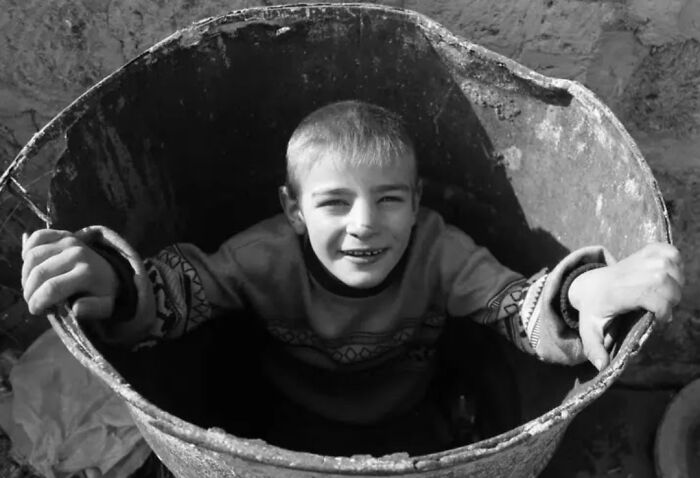 A child smiling from inside a large metal container, capturing the innocence and resilience of childhood in black and white photography.