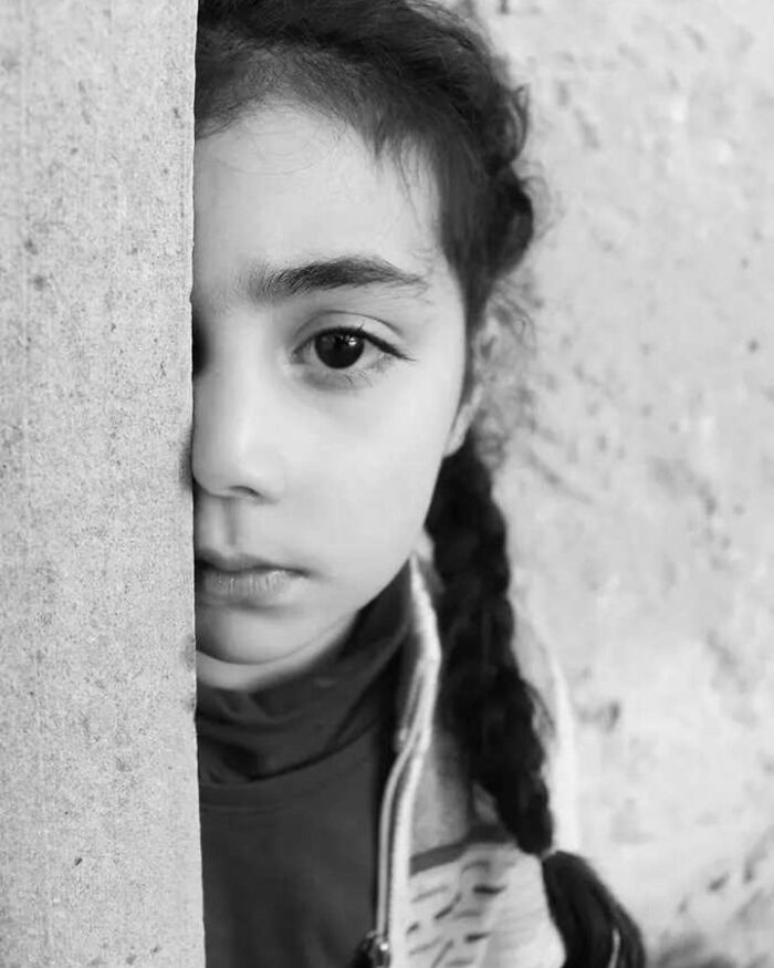 Black and white photo of a child with braided hair, embodying innocence and resilience, standing against a textured wall.