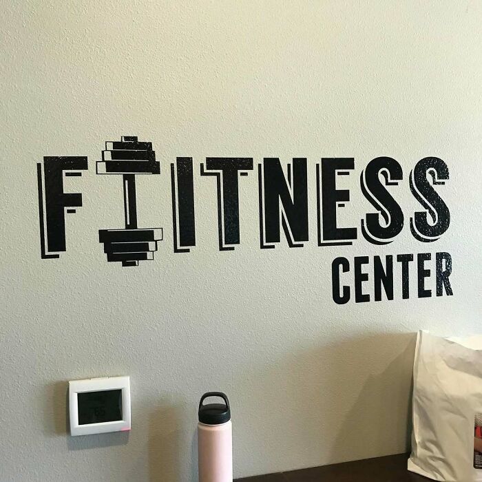 Wall with "Fitness Center" sign where dumbbell replaces the letter "I," highlighting a design fail.