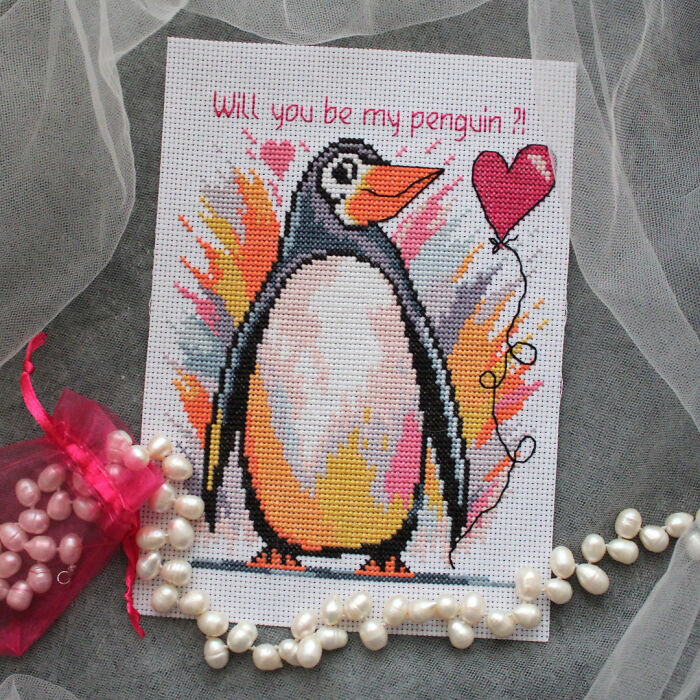 Penguin cross stitch pattern with a heart balloon and the text "Will you be my penguin?" on a fabric background.