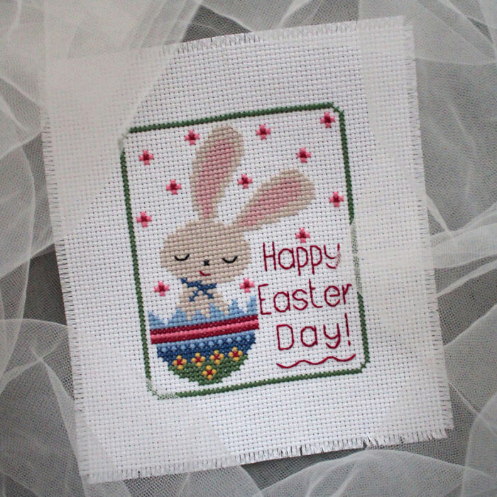 Cross stitch pattern of a bunny in an egg with text "Happy Easter Day" on fabric.