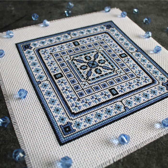 Intricate digital cross stitch pattern in blue and black with geometric design, surrounded by small blue beads.