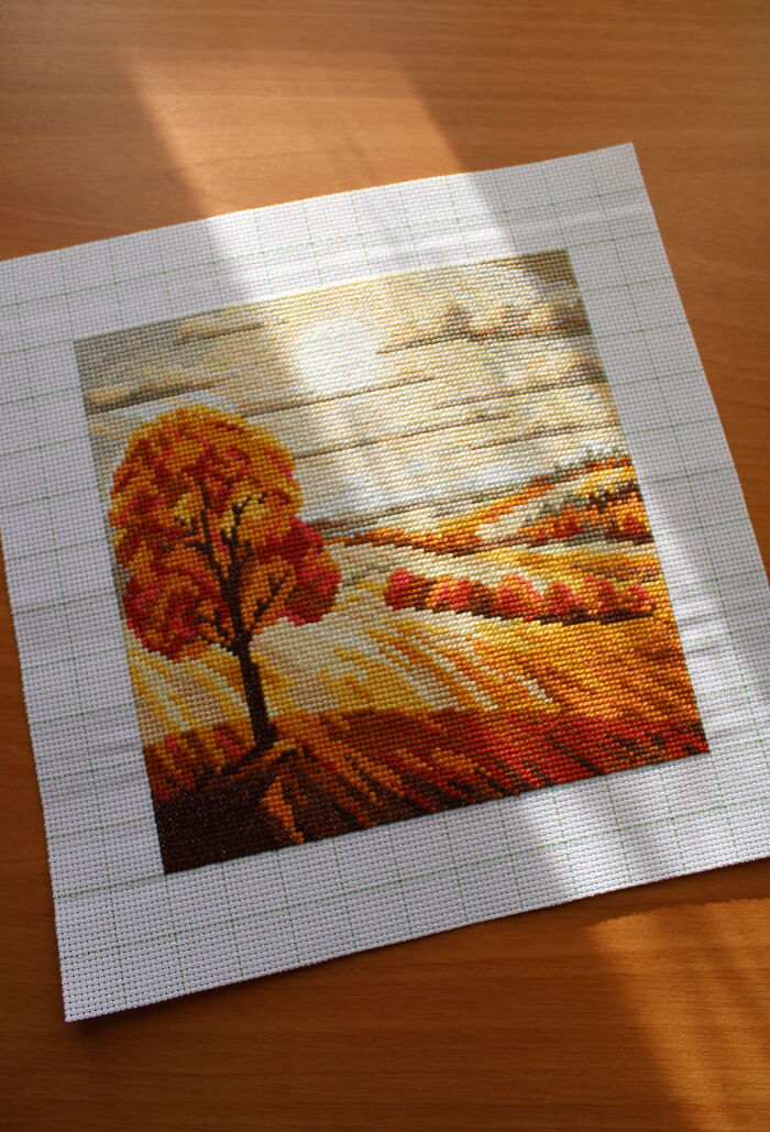 Digital cross stitch pattern of an autumn landscape with a tree, sunlight casting shadows on a wooden surface.