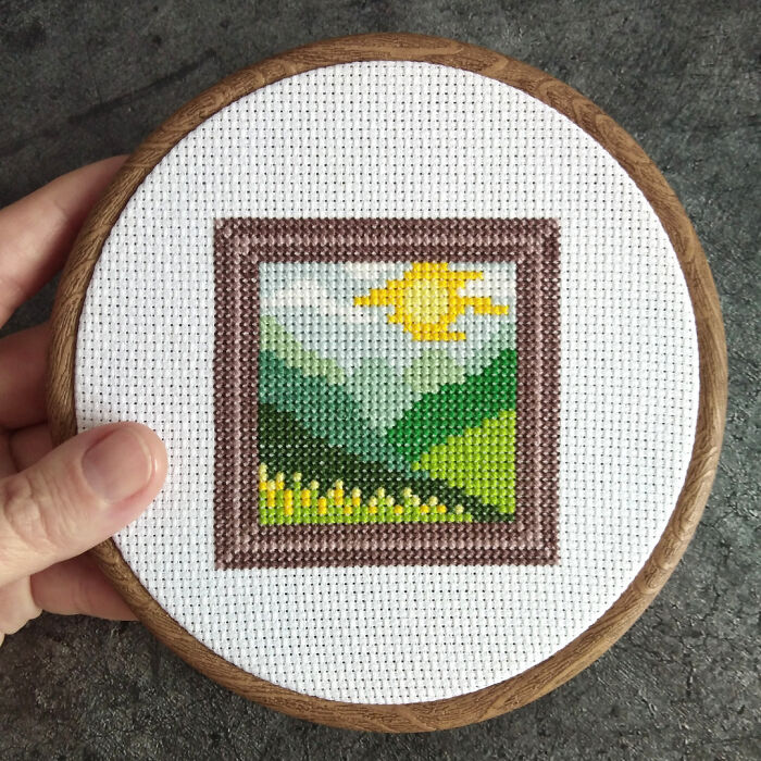 Cross stitch pattern with a sunny landscape in a circular frame, held by hand.