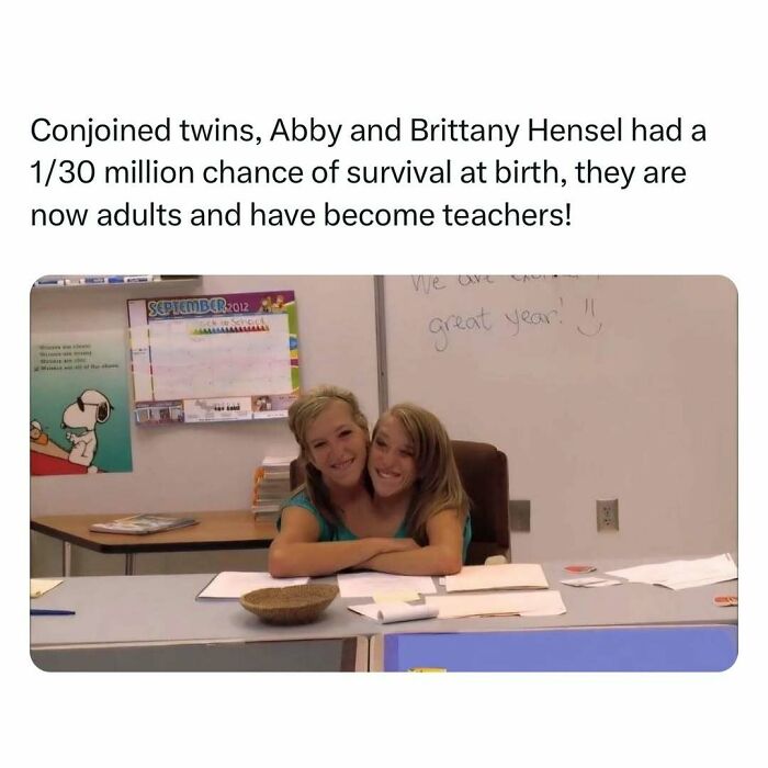 Conjoined twins sitting at a classroom desk, smiling joyfully; they are now teachers, bringing heartwarming inspiration.