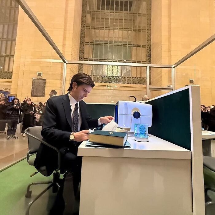 A man in a suit works at a desk in a glass box. Crowds watch, illustrating the strange nature of the internet.