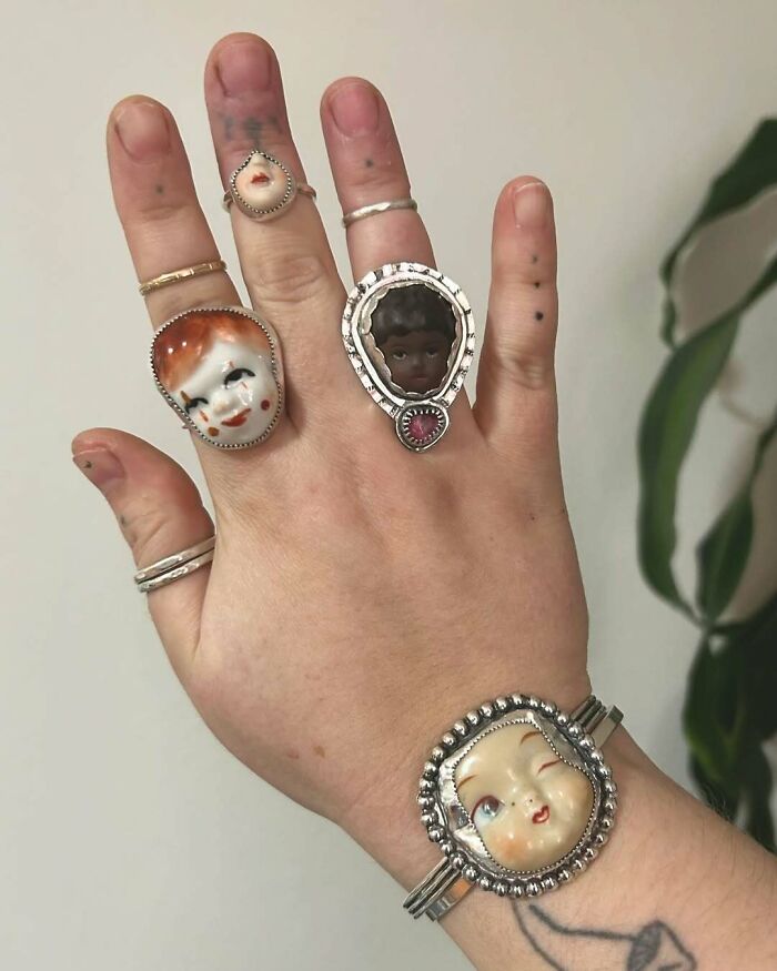 A hand wearing interesting creepy handmade crafts, including unique doll face rings.