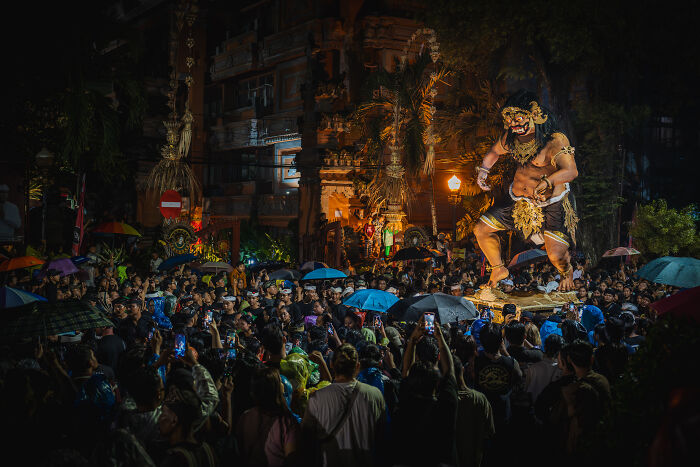 Crowd gathered under umbrellas at night, watching a large statue during Bali's Galungan celebration.