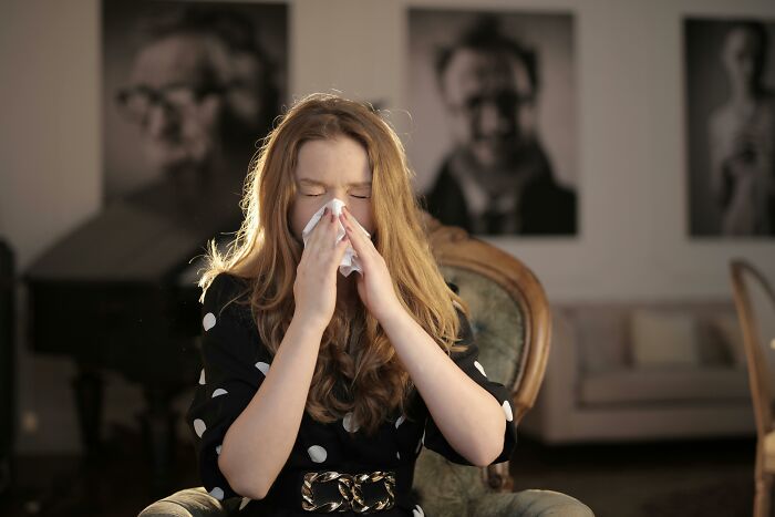 Woman in polka dot dress sneezing, possibly illustrating superstitions, sitting on a vintage chair in a living room.