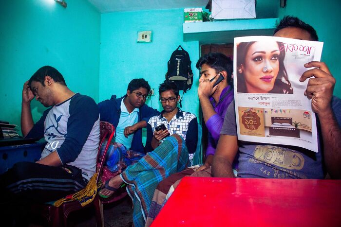 Street photography by Md Enamul Kabir featuring men indoors, one reading a newspaper, another on the phone, in a colorful room.