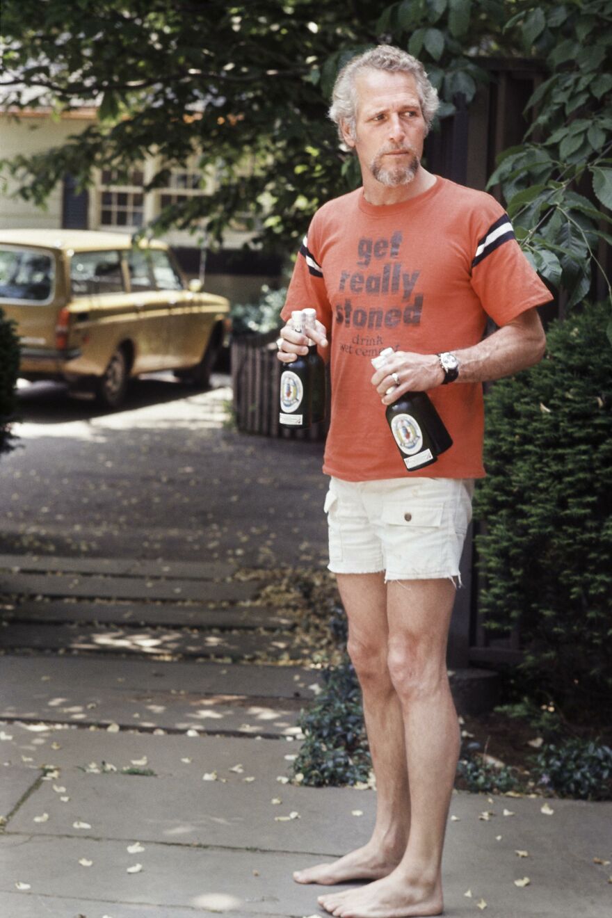 Barefoot man holding beers, wearing shorts and a vintage shirt, standing on a leafy sidewalk. Historical photo.
